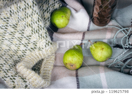 Knitted sweater, three pear fruits and thermos mug on checkered blanket in open car trunk. Autumn still life. Road trip picnic. Travel lifestyle moments. Fruits and coffee mug in trunk car.  119625798