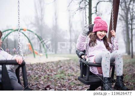 one smiling girl sitting on the swing at children playground on a foggy day, playing with the friend 119626477