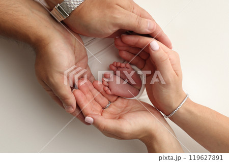 The palms of the parents. A father and mother hold the feet of a newborn child in a white blanket on a white background.. The feet of a newborn in the hands of parents. Photo of foot, heels and toes 119627891