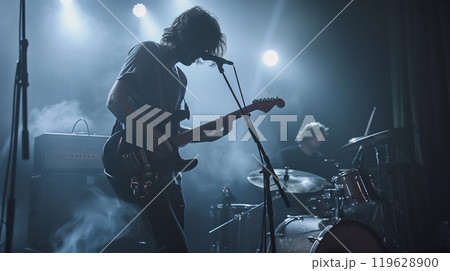 A guitarist passionately plays while a drummer keeps the rhythm, surrounded by dim lights and smoke in an intimate concert setting 119628900