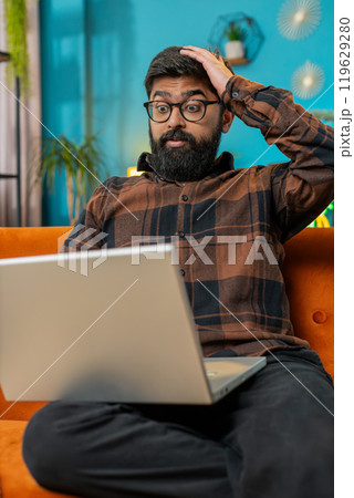 Upset Indian man sitting on home sofa using laptop having anxiety stress loss bad fail news virus Upset Indian man sitting on home sofa using laptop having anxiety stress loss bad fail news virus 119629280
