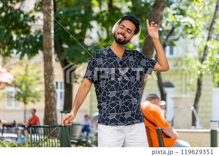 Happy relaxed young Indian man in wireless headphones listening disco music dancing outdoors Happy relaxed young Indian man in wireless headphones listening disco music dancing outdoors 119629351
