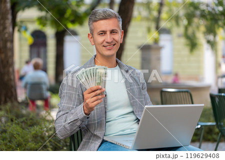Happy rich man using laptop winner victory holding cash money in dollar banknotes sitting on chair 119629408