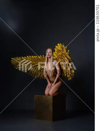 Model poses gracefully with golden wings on a pedestal in a dark studio setting Model poses gracefully with golden wings on a pedestal in a dark studio setting 119630701