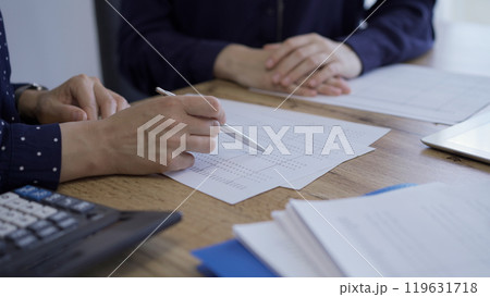 Accountants or auditors reviewing financial documents while sitting at the wooden desk in office. Professionals analyzing paper reports and using calculators, side view. Audit and taxes Accountants or auditors reviewing financial documents while sitting at the wooden desk in office. Professionals analyzing paper reports and using calculators, side view. Audit and taxes 119631718