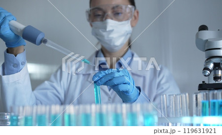 Scientist woman, wearing a lab coat, mask, blue gloves and safety glasses, is using a micropipette to drop a liquid into a test tubes. Medicine and science Scientist woman, wearing a lab coat, mask, blue gloves and safety glasses, is using a micropipette to drop a liquid into a test tubes. Medicine and science 119631921