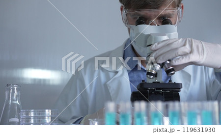 Male scientist, wearing a lab coat, mask, white gloves and safety glasses, is using a binocular microscope surrounded by test tubes in laboratory Male scientist, wearing a lab coat, mask, white gloves and safety glasses, is using a binocular microscope surrounded by test tubes in laboratory 119631930