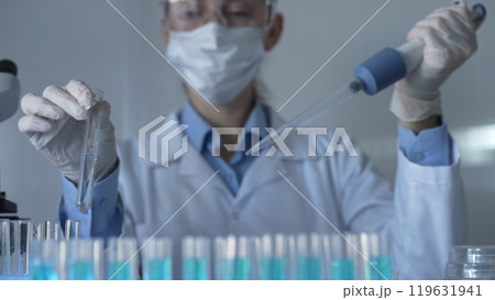 Scientist woman, wearing a lab coat, mask, blue gloves and safety glasses, is using a micropipette to drop a liquid into a test tubes. Medicine and science Scientist woman, wearing a lab coat, mask, blue gloves and safety glasses, is using a micropipette to drop a liquid into a test tubes. Medicine and science 119631941