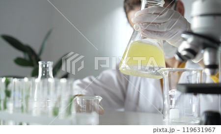Woman scientist researcher wearing white protective gloves is shaking up a yellow chemical solution inside an Erlenmeyer flask in a laboratory setting, portrait view. Science and medicine 119631967
