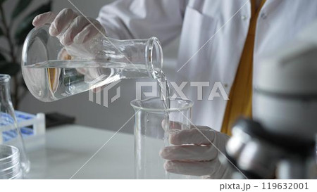Unknown scientist with a lab coat and white protective gloves is carefully pouring a liquid from a beaker into an Erlenmeyer flask, close up. Science and medicine concept 119632001