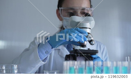 Scientist woman, wearing a lab coat, mask, blue gloves and safety glasses, is adjusting a microscope surrounded by test tubes in laboratory. Medicine and science concept 119632003