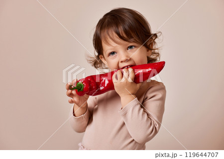 happy child girl bites fresh red pepper on beige background. healthy baby food. kid loves vegetables 119644707