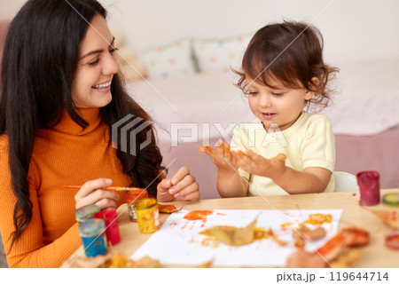 happy little child girl and mom spending good time painting hands and autumn yellow leaves together happy little child girl and mom spending good time painting hands and autumn yellow leaves together 119644714