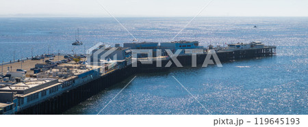 The Santa Cruz Wharf stretches into the Pacific Ocean, with buildings and parked cars along its length. Boats are visible in the distance under sunlight. The Santa Cruz Wharf stretches into the Pacific Ocean, with buildings and parked cars along its length. Boats are visible in the distance under sunlight. 119645193