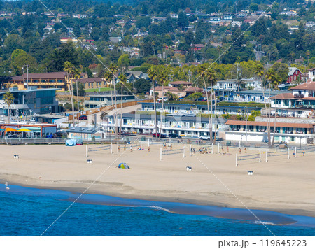 Aerial view of Santa Cruz, California, featuring a sandy beach with volleyball courts, palm trees, low rise buildings, and the ocean with gentle waves. 119645223