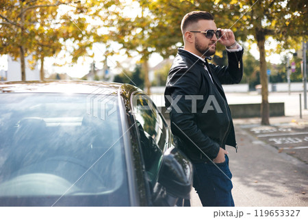 Portrait of a businessman standing beside his luxury car, placing his left hand on his sunglasses 119653327