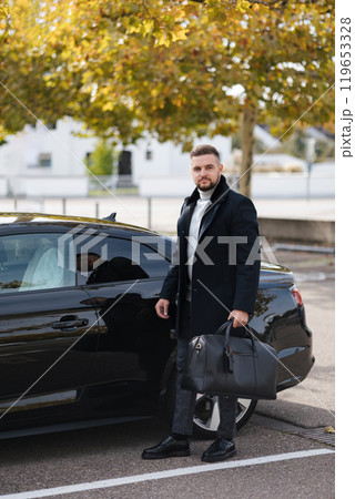 Businessman take a bag from his luxury car and go to airport. Stylish bearded man in coat on auto parking Businessman take a bag from his luxury car and go to airport. Stylish bearded man in coat on auto parking 119653328