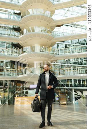 Man in coat walking though the big office. Guy hold travel bag. Stylish guy after working day 119653368