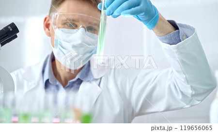 Scientist with protective gloves and glasses examining lab tube with a green fluid. Microbiology and science concept Scientist with protective gloves and glasses examining lab tube with a green fluid. Microbiology and science concept 119656065