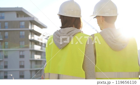 Man and woman engineers wearing safety hard hats and vests holding blueprint and discussing something on construction site at sunrise, back view. Architecture and engineering concept 119656110