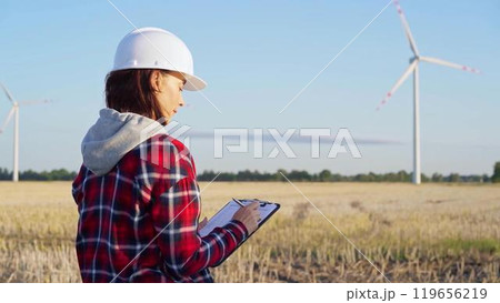 Woman engineer wearing a white protective helmet is taking notes with a clipboard in a field with wind turbines, as the sun sets. Clean energy and engineering concept Woman engineer wearing a white protective helmet is taking notes with a clipboard in a field with wind turbines, as the sun sets. Clean energy and engineering concept 119656219