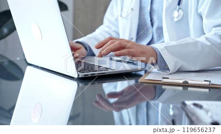 Doctor man with blue shirt and medical coat is using laptop computer in clinic office while sitting at the glass desk. Medicine concept 119656237
