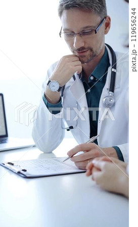 Doctor and a patient. The physician, wearing a white medical coat over a green shirt, gesturing with his hands during a consultation in the clinic. Medicine concept Doctor and a patient. The physician, wearing a white medical coat over a green shirt, gesturing with his hands during a consultation in the clinic. Medicine concept 119656244