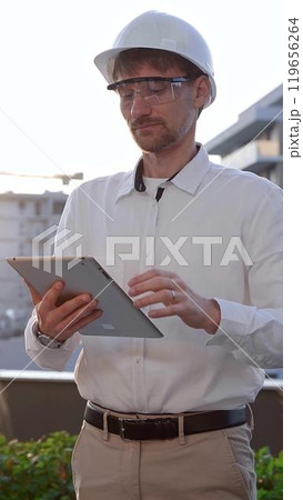 Male architectural engineer, wearing a white shirt and hard hat, is using a digital tablet while inspecting a building site at sunrise, front and vertical view. Construction and engineering 119656264