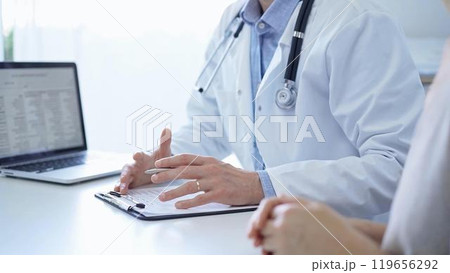 A doctor and a patient. The physician, wearing a white medical coat over a blue shirt, is gesturing with his hands during a consultation in the clinic. Medicine A doctor and a patient. The physician, wearing a white medical coat over a blue shirt, is gesturing with his hands during a consultation in the clinic. Medicine 119656292