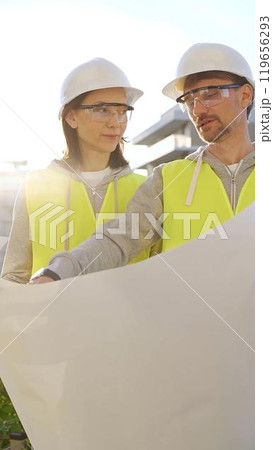 Two architects wearing safety hard hats and vests holding blueprint and discussing something on construction site at sunrise, front vertical view. Architecture engineering and teamwork concept Two architects wearing safety hard hats and vests holding blueprint and discussing something on construction site at sunrise, front vertical view. Architecture engineering and teamwork concept 119656293