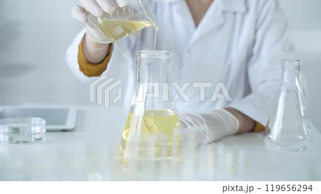 A scientist, wearing a lab coat and white protective gloves, is pouring a yellow oily liquid from one beaker to another in laboratory, close up. Medicine and science concept 119656294