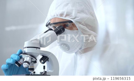 Scientist in ppe analyzing samples using a microscope in a laboratory setting. Close-up of a focused researcher in a lab coat mask and protective gear Scientist in ppe analyzing samples using a microscope in a laboratory setting. Close-up of a focused researcher in a lab coat mask and protective gear 119656315