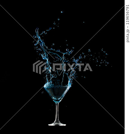 Pouring Clear Cocktail glass with spilled blue liquid in air. Blue soda splashing out of glass with curve spill and splashing. Black background isolated element 119656791