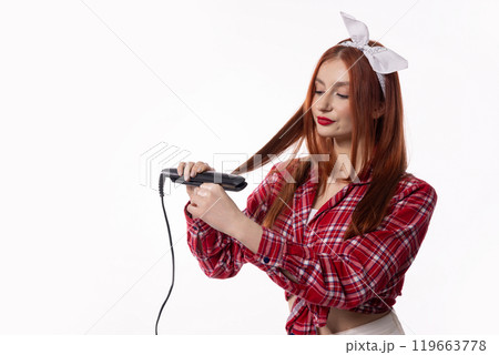 A Woman is Styling Her Hair with a Straightener While Wearing a Fashionable and Trendy Outfit A Woman is Styling Her Hair with a Straightener While Wearing a Fashionable and Trendy Outfit 119663778