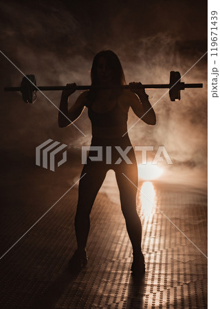 A powerful silhouette of a determined female weightlifter in a dynamic gym symbolizes athleticism, courage, and perseverance, embodying the spirit of a warrior pushing boundaries and defying limits 119671439