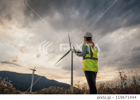 A dedicated female technician is carefully inspecting a wind turbine during a stunning sunset, set against the backdrop of a beautiful rural landscape filled with natures wonders A dedicated female technician is carefully inspecting a wind turbine during a stunning sunset, set against the backdrop of a beautiful rural landscape filled with natures wonders 119671444