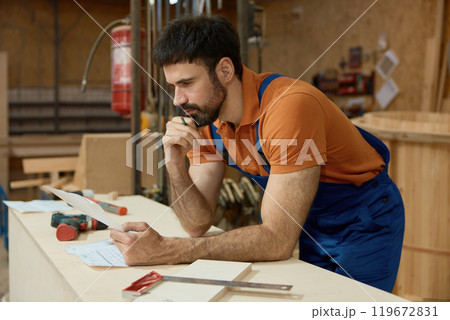 Pensive man carpenter reading blueprints and thinking during woodworking process Pensive man carpenter reading blueprints and thinking during woodworking process 119672831
