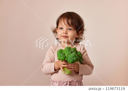 child girl holding fresh green raw broccoli on beige background. healthy baby food. kid loves vegetables 119673119