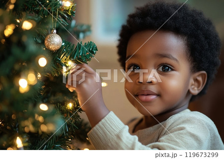 Adorable little African American boy with curly hair decorating Christmas tree with golden balls in living room at home, child toddler preparing to celebrate new year 119673299