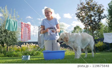 A woman wrings out laundry outdoors with a Golden Retriever standing nearby on a sunny day. 119675648