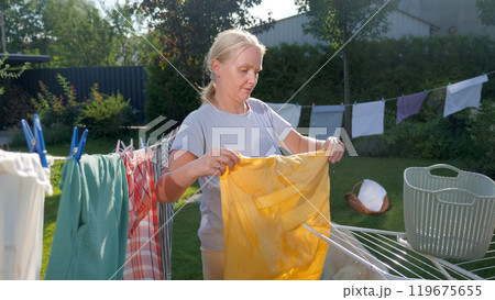 A woman hangs a yellow garment to dry outdoors on a sunny day, surrounded by colorful clothes on a line in a peaceful backyard setting. 119675655