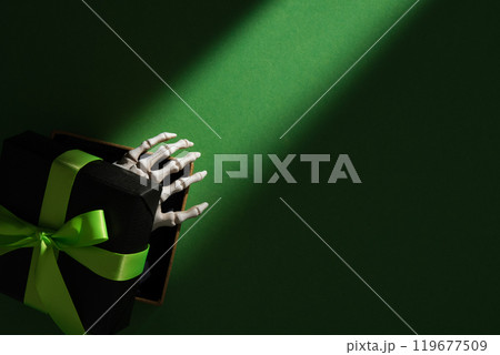 Skeleton hand emerging from black gift box with green ribbon on dark green background, mysterious Halloween present concept. Copyspace. 119677509