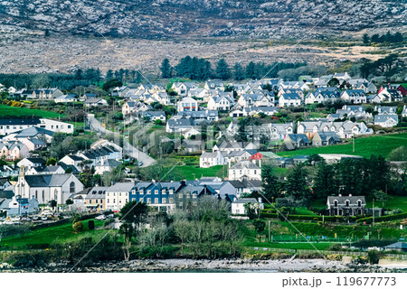 A coastal town with a diverse array of houses, a church, and a small harbor in the foreground. The town is surrounded by lush greenery and a rocky hill in the background. 119677773