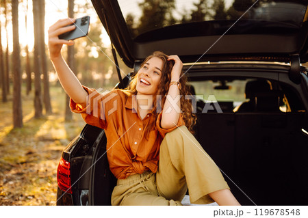 Happy stylish female tourist in trunk of a car with phone, taking a selfie, talking on a video call. 119678548
