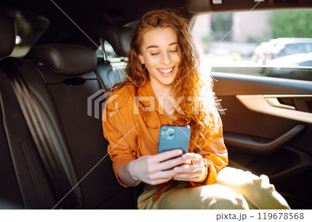 Cheerful woman with curly hair and stylish clothes sits in back seat of a car, uses a mobile phone. Cheerful woman with curly hair and stylish clothes sits in back seat of a car, uses a mobile phone. 119678568