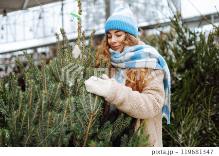 Smiling woman in warm clothes chooses a Christmas tree at the market. Holiday concept. Smiling woman in warm clothes chooses a Christmas tree at the market. Holiday concept. 119681347