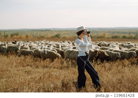 Young woman with a camera in front of a herd of sheep. Nature, fashion, vacation and lifestyle. 119681428