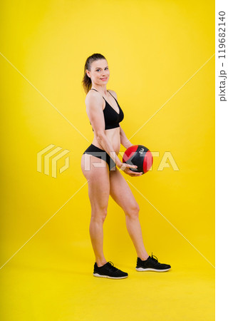Sportive caucasian cross fit woman with fitness ball, look at camera. Studio dark yellow background 119682140