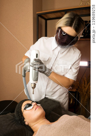 Cosmetologist wearing protective eyewear conducts facial treatment 119689258