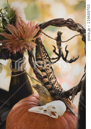 Atmospheric autumn altar. Dry birds feather, dry claws and skull, pumpkin, willow wreath, beautiful long-term dahlia flowers, vertical format. Atmospheric autumn altar. Dry birds feather, dry claws and skull, pumpkin, willow wreath, beautiful long-term dahlia flowers, vertical format. 119689703